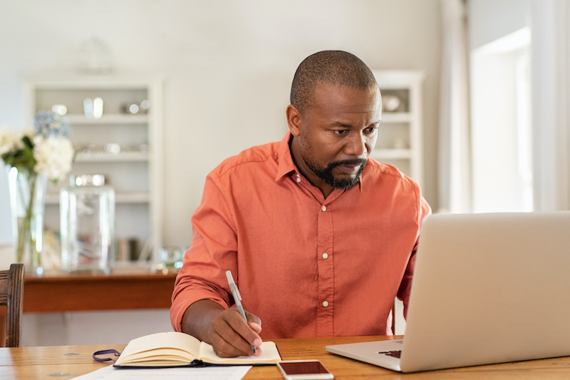 african american man taking notes next to his laptop at home