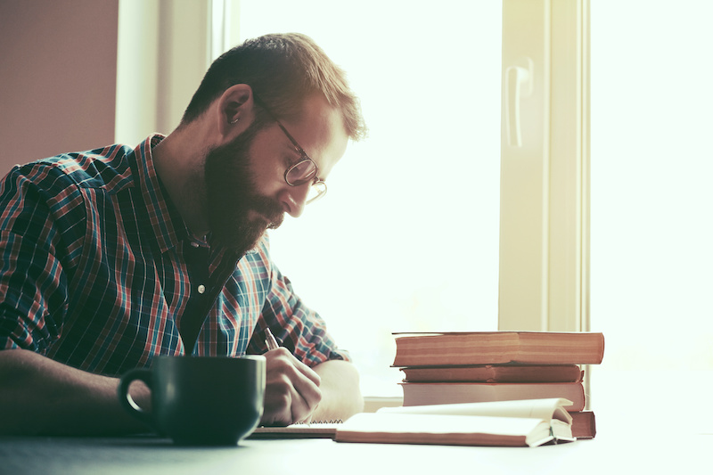 bearded man taking notes next to a stack of books