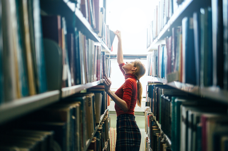 woman reaching up high to grab a book in a library