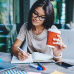 Black haired woman taking notes in front of laptop with a coffee in her hand