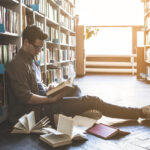 Man on the floor of a library with books all around him, searching for a gap in literature