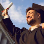 Ph.D. graduate standing near university holding up diploma