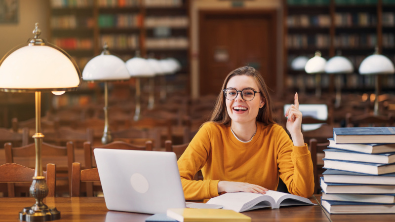 smiling woman with glasses sitting at a table in the library with a laptop and books