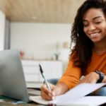 Smiling young black woman working on laptop and writing in notebook