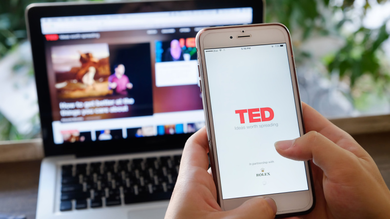 person holding a phone in front of a laptop with the ted talk site on